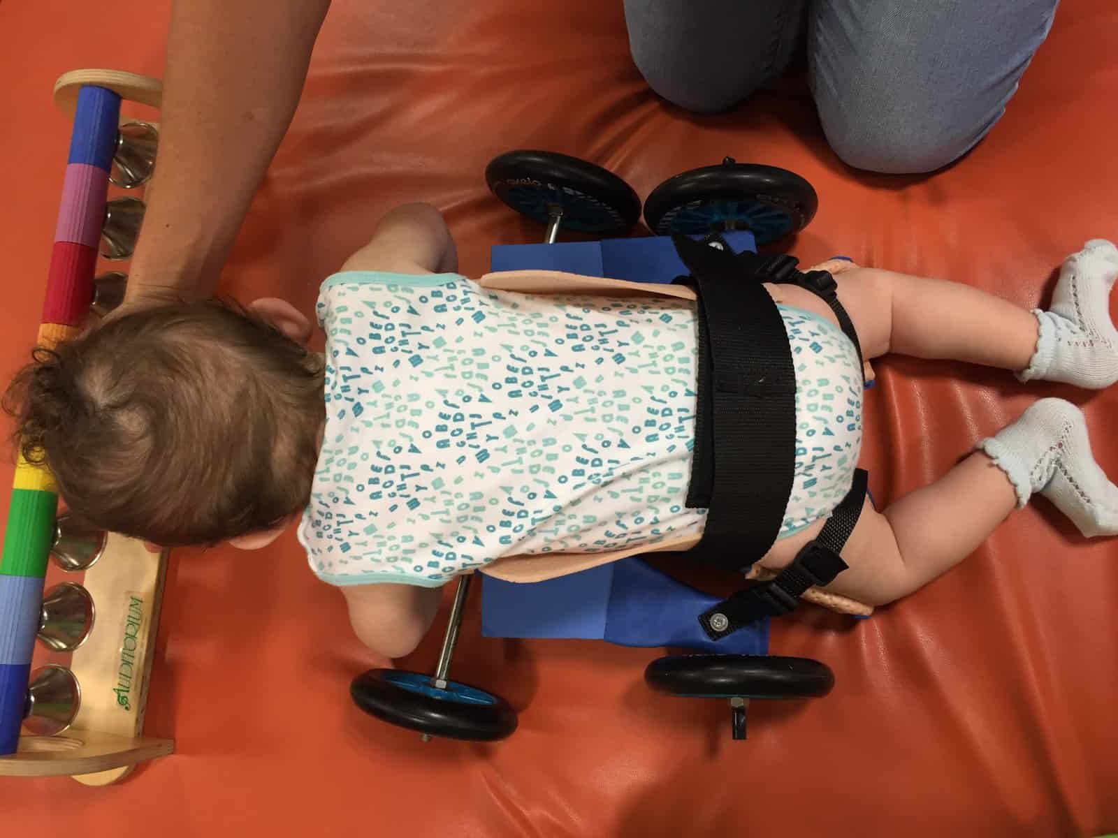 A baby lying face down on a custom floor scooter, secured with straps, reaches for a toy