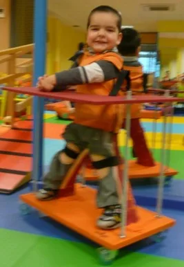 A boy standing in his orange stander with a support hoop, in the therapy room