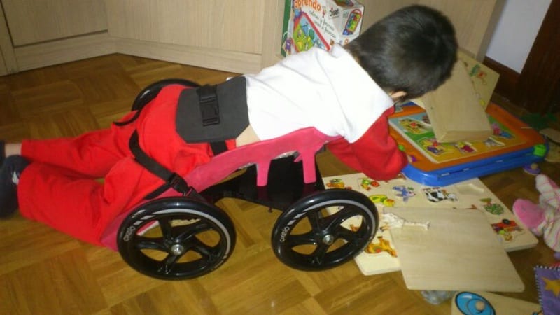 A child lying face down on a custom floor scooter plays with puzzles on the floor at home