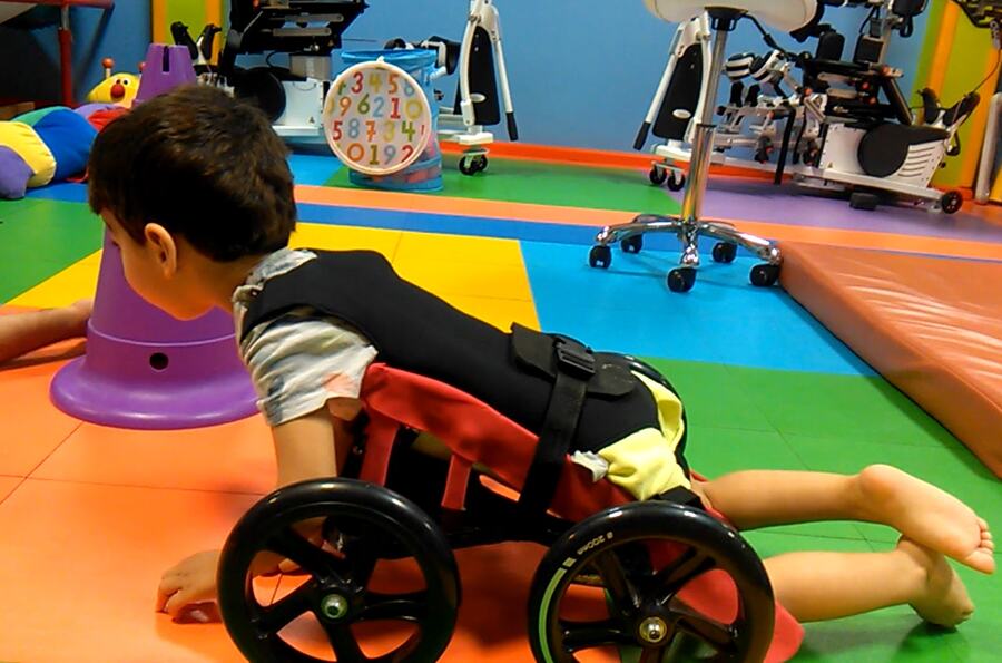 A child lying face down on a custom floor scooter pushes along with his arms in the clinic room