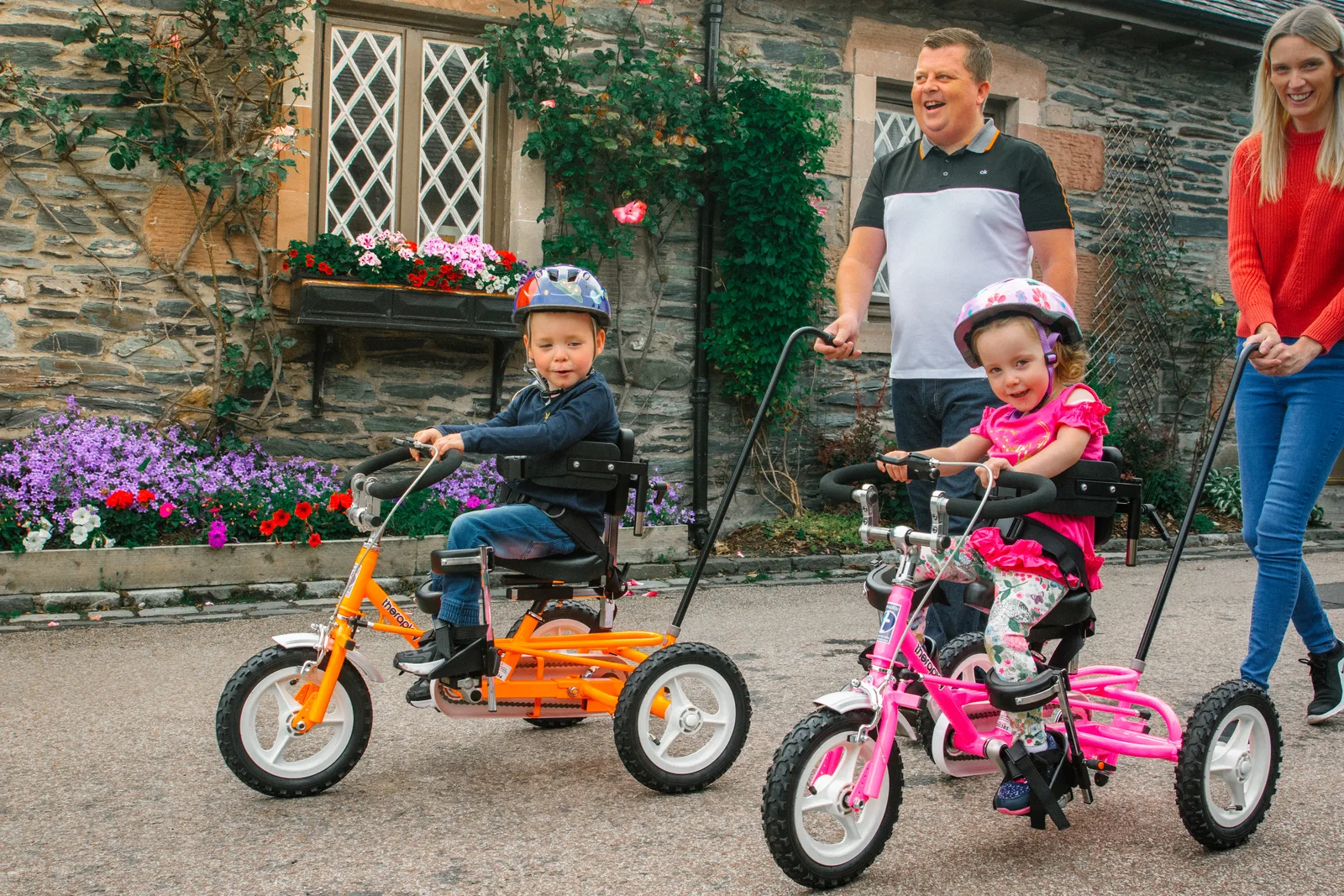 Two children pedal their adapted tricycles alongside two adults outside a house