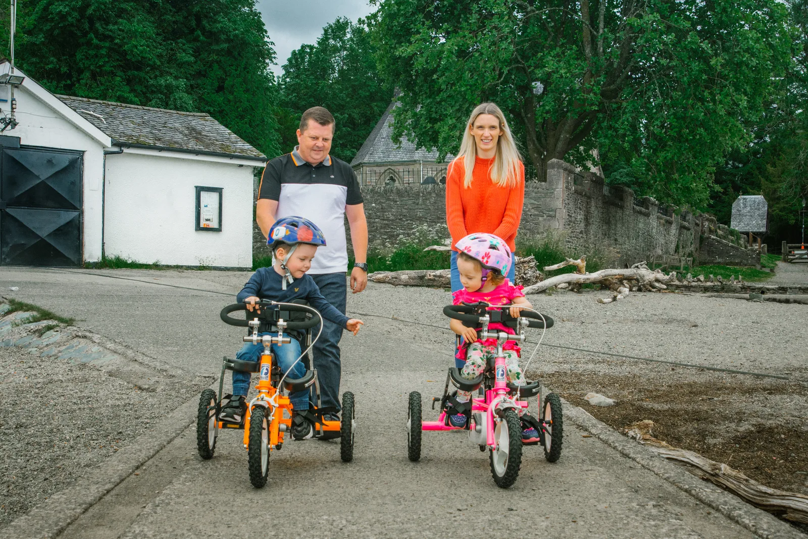 Two children in helmets pedal along a path on their adapted tricycles