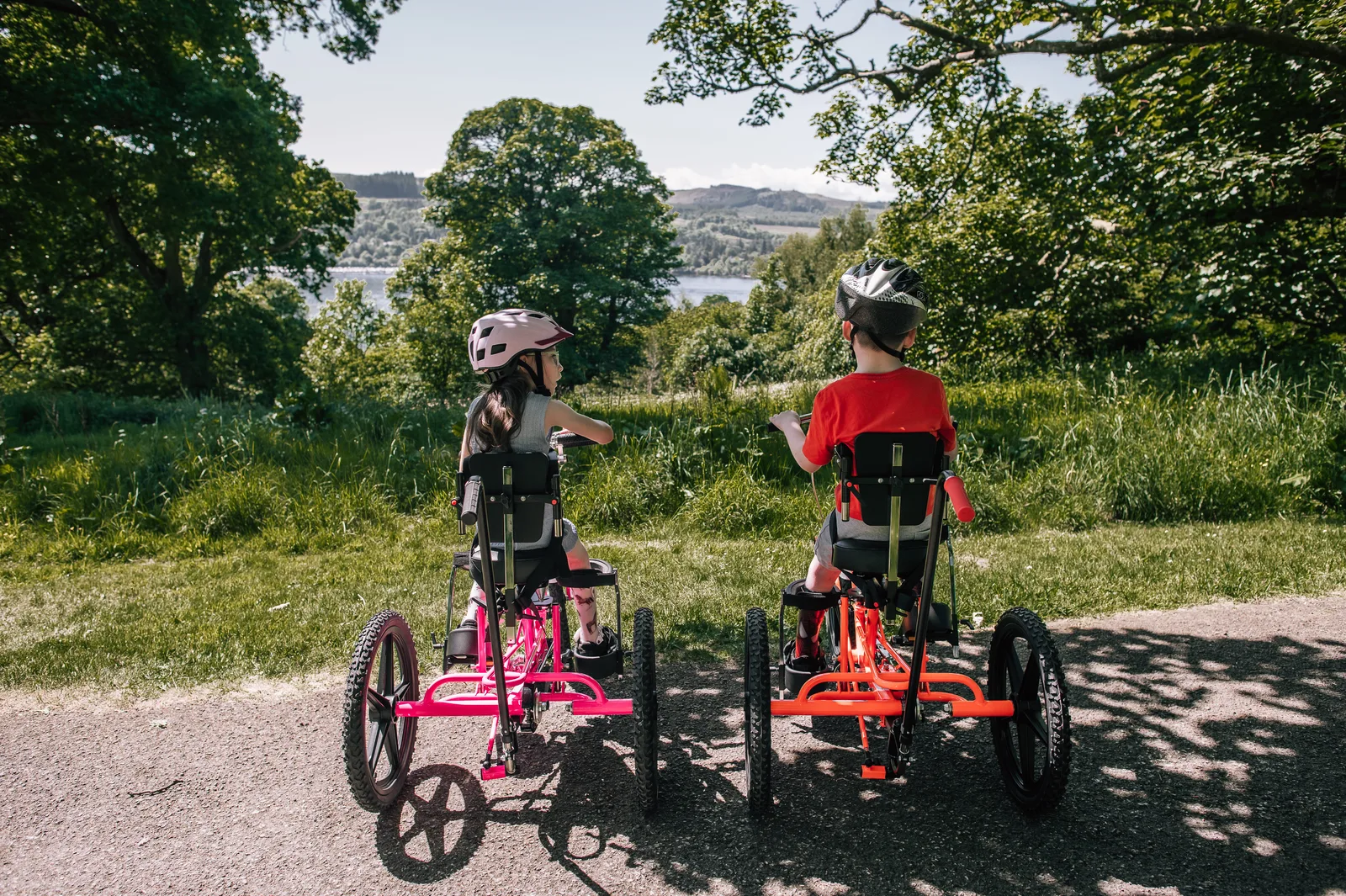 Two children in helmets ride along a country path on adapted tricycles