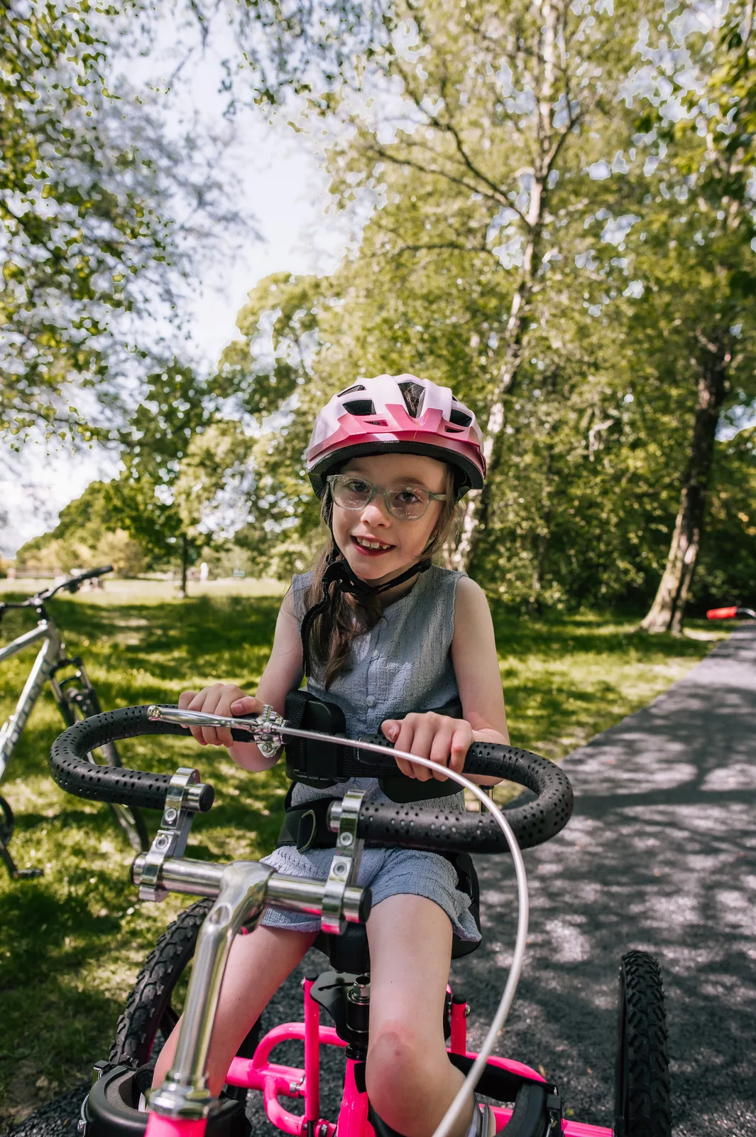 A girl in a pink helmet pedals her adapted tricycle along a tree-lined path